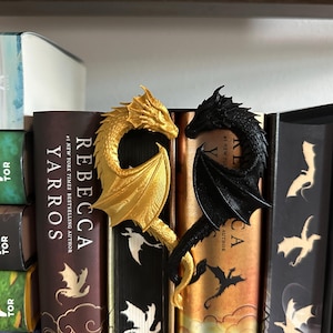 May include: Two dragon bookends, one gold and one black, are displayed on a bookshelf with several books. The bookends are in the shape of dragons with their wings spread out.
