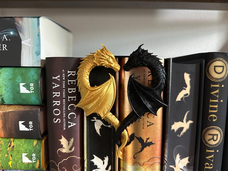 May include: Two dragon bookends, one gold and one black, are displayed on a bookshelf with several books. The bookends are in the shape of dragons with their wings spread out.