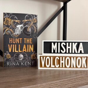 Puede incluir: Un libro titulado "Hunt the Villain" con un dise&ntilde;o de calavera y serpiente, y dos letreros decorativos con los nombres "Mishka Volchonok". Los art&iacute;culos se exhiben en una estanter&iacute;a de madera.