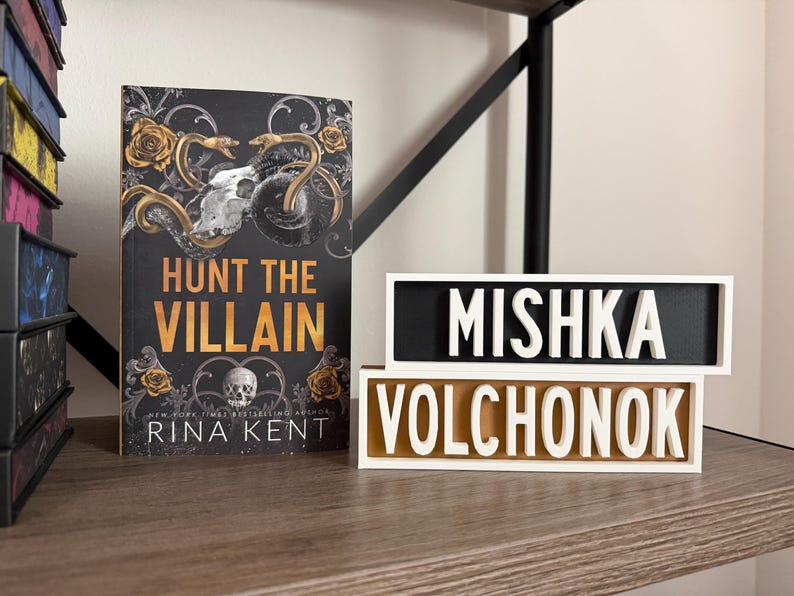 Puede incluir: Un libro titulado "Hunt the Villain" con un dise&ntilde;o de calavera y serpiente, y dos letreros decorativos con los nombres "Mishka Volchonok". Los art&iacute;culos se exhiben en una estanter&iacute;a de madera.