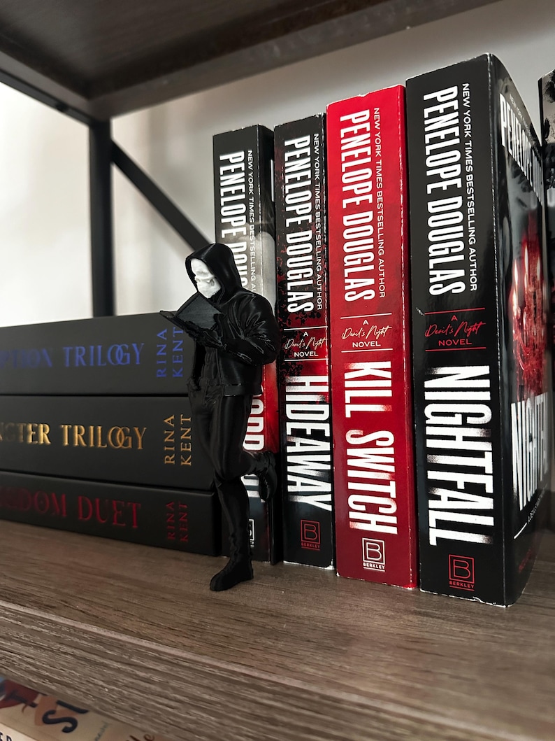 Puede incluir: Una figura negra de una figura encapuchada leyendo un libro, colocada frente a una estanter&iacute;a de libros. Los libros incluyen t&iacute;tulos como "Hideaway", "Kill Switch" y "Nightfall" de Penelope Douglas. La estanter&iacute;a es de madera.