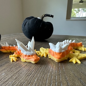 Puede incluir: Dos figuritas de dragón de color maíz dulce con detalles blancos y amarillos. Los dragones están tumbados sobre una superficie de madera.