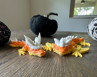 May include: Two candy corn-colored dragon figurines with white and yellow accents. The dragons are lying on a wooden surface.