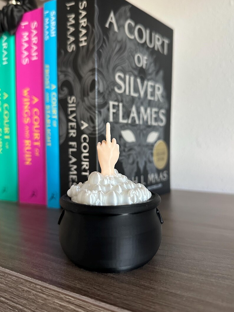Op de afbeelding: Een zwarte ketel met wit schuim dat eroverheen borrelt. Een middelvinger steekt uit het schuim. De ketel staat op een houten oppervlak voor een boekenkast met verschillende boeken getiteld "A Court of Silver Flames" van Sarah J. Maas.