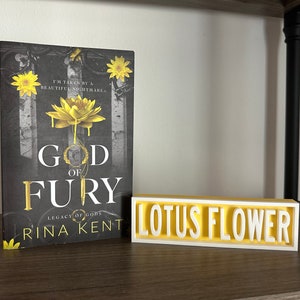 Może przedstawiać: Książka zatytułowana "God of Fury" autorstwa Riny Kent, z czarno-złotą okładką z ż&oacute;łtym kwiatem lotosu. Obok biały i ż&oacute;łty znak "Lotus Flower".