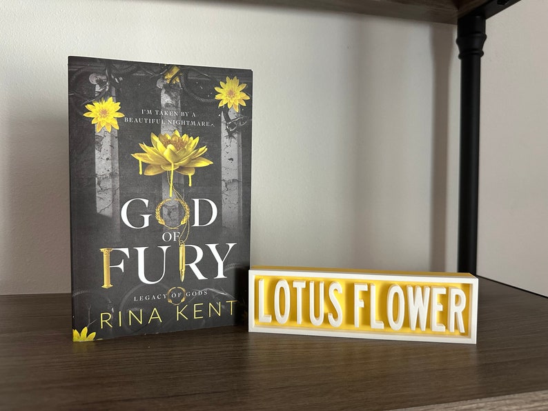 Może przedstawiać: Książka zatytułowana "God of Fury" autorstwa Riny Kent, z czarno-złotą okładką z ż&oacute;łtym kwiatem lotosu. Obok biały i ż&oacute;łty znak "Lotus Flower".