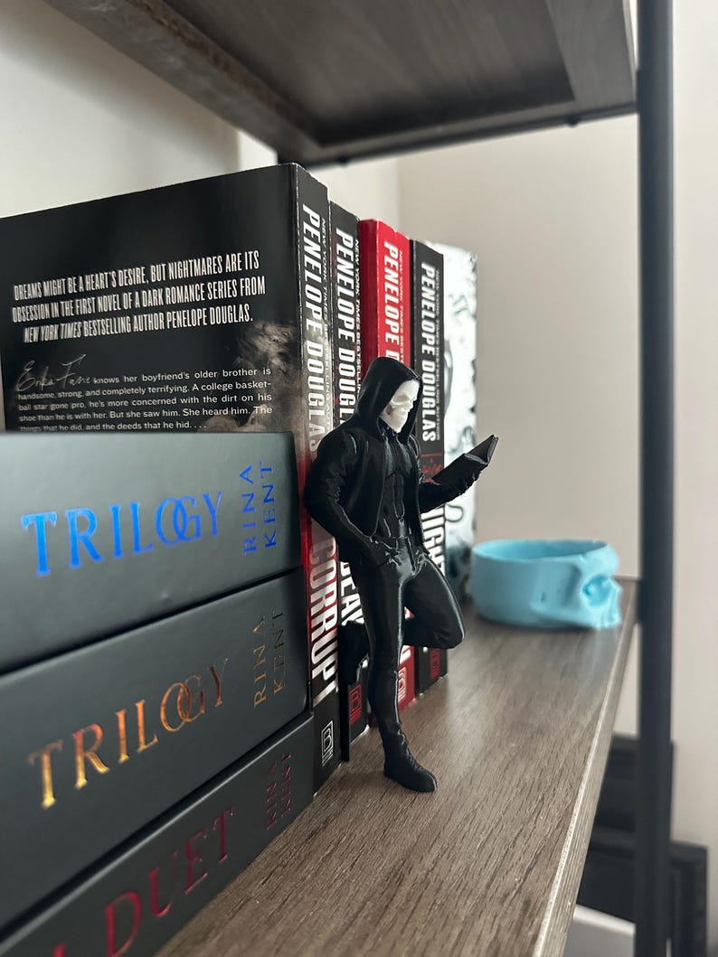 Puede incluir: Una figura negra de un personaje con capucha, apoyada en una pila de libros. Los libros tienen t&iacute;tulos como "Trilogy" y "Corrupt" y est&aacute;n en una estanter&iacute;a de madera. Un recipiente azul con forma de calavera tambi&eacute;n est&aacute; en la estanter&iacute;a.