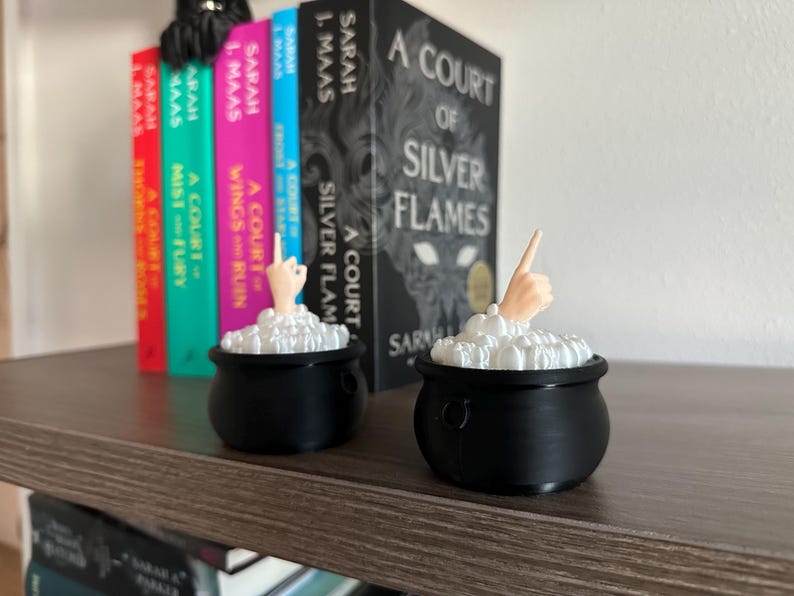 Op de afbeelding: Twee zwarte, ketelvormige containers met wit schuim en een hand die uit elk uitsteekt. De ketels staan op een houten plank met een rij boeken erachter. De boeken hebben titels als "A Court of Silver Flames" en "A Court of Wings and Ruin".
