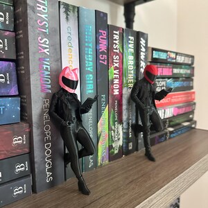 May include: A bookshelf with several books and two black figurines. The books have titles like "TRYST SIX VENOM" and "PUNK 57" by Penelope Douglas. The figurines are wearing helmets and appear to be reading.
