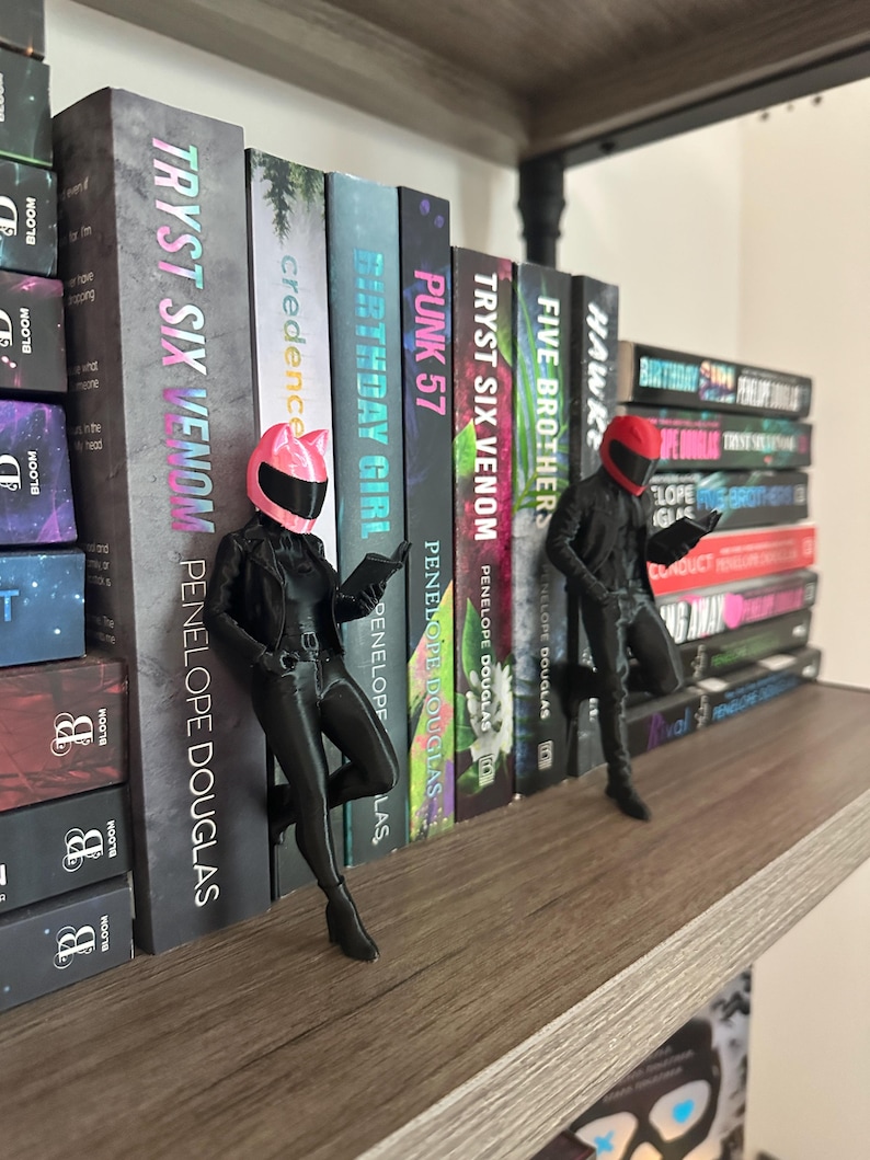 May include: A bookshelf with several books and two black figurines. The books have titles like "TRYST SIX VENOM" and "PUNK 57" by Penelope Douglas. The figurines are wearing helmets and appear to be reading.