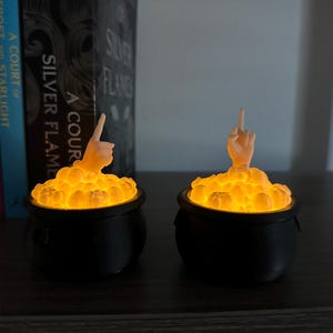Op de afbeelding: Twee zwarte, ketelvormige kaarsenhouders met een gloeiend oranje licht erin. Elke ketel heeft een klein, wit, handvormig figuurtje dat uit de bovenkant steekt.