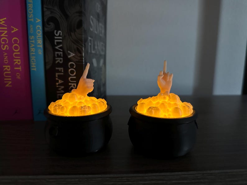 Op de afbeelding: Twee zwarte, ketelvormige kaarsenhouders met een gloeiend oranje licht erin. Elke ketel heeft een klein, wit, handvormig figuurtje dat uit de bovenkant steekt.