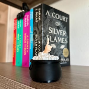 Op de afbeelding: Een zwarte ketel met een witte hand die uit de bovenkant steekt, omringd door wit schuim. De ketel staat op een houten plank met een rij boeken erachter. De boeken behoren allemaal tot de serie "A Court of Thorns and Roses" van Sarah J. Maas.