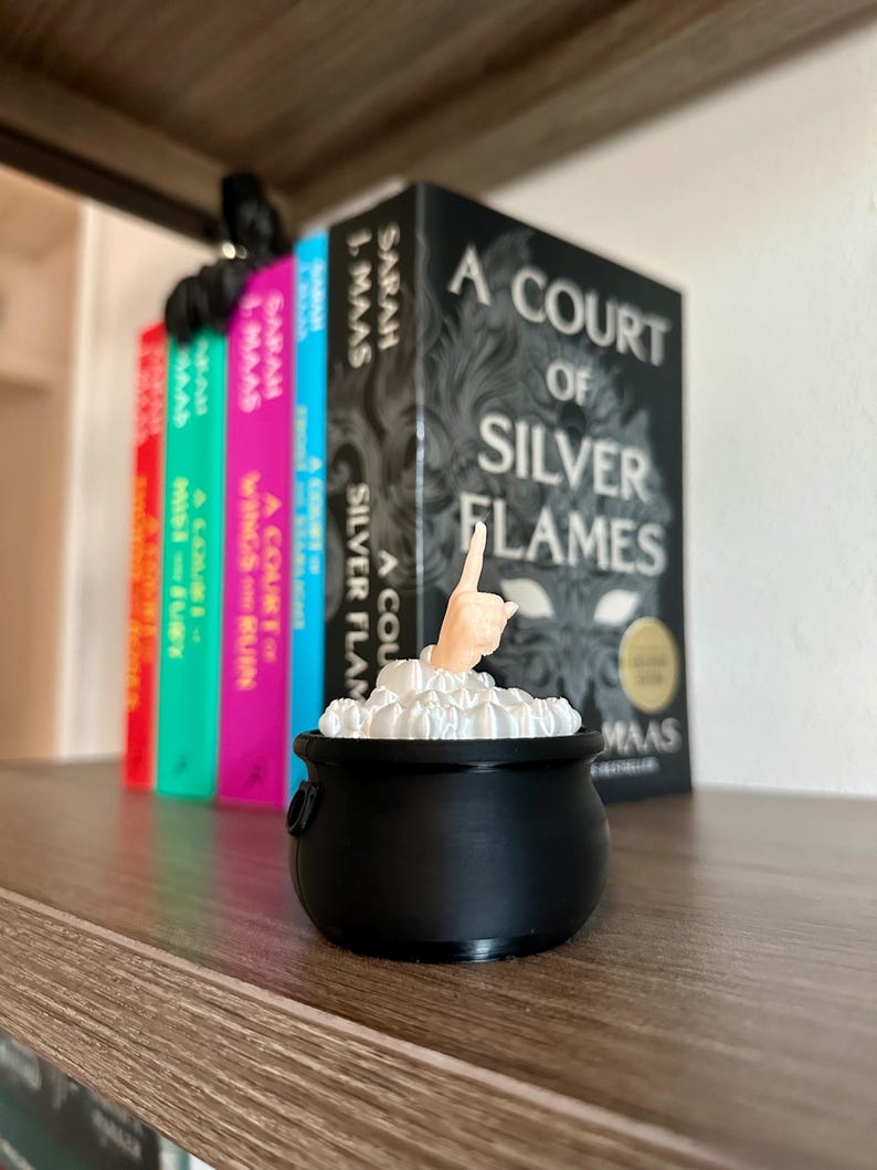 Op de afbeelding: Een zwarte ketel met een witte hand die uit de bovenkant steekt, omringd door wit schuim. De ketel staat op een houten plank met een rij boeken erachter. De boeken behoren allemaal tot de serie "A Court of Thorns and Roses" van Sarah J. Maas.