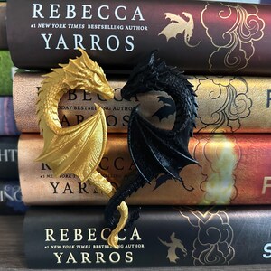 May include: A pair of dragon bookends, one gold and one black, are displayed on a stack of books. The books are all by the same author, Rebecca Yarros, and feature different titles from her "Four Winds" series.