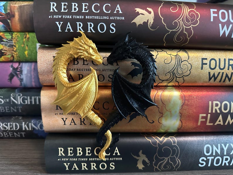May include: A pair of dragon bookends, one gold and one black, are displayed on a stack of books. The books are all by the same author, Rebecca Yarros, and feature different titles from her "Four Winds" series.