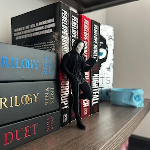 Puede incluir: Una estanter&iacute;a de madera exhibe varios libros con lomos negros y rojos. Una figura de un personaje con un atuendo negro con capucha y rostro de calavera est&aacute; cerca de los libros. Los libros incluyen t&iacute;tulos como "Trilogy" y "Duet".