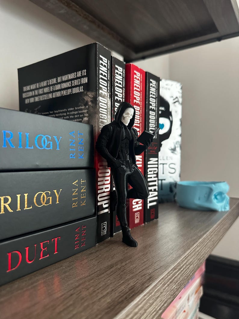 Puede incluir: Una estanter&iacute;a de madera exhibe varios libros con lomos negros y rojos. Una figura de un personaje con un atuendo negro con capucha y rostro de calavera est&aacute; cerca de los libros. Los libros incluyen t&iacute;tulos como "Trilogy" y "Duet".