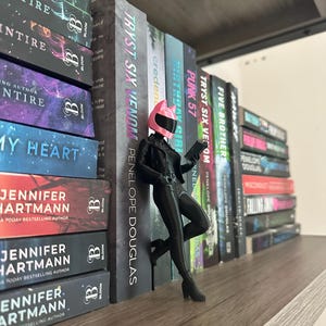 May include: A shelf displays a collection of books with titles like "TRYST SIX VENOM" and "PUNK 57." A figurine in a black outfit and pink helmet leans against the books. The books have colorful covers and the "BLOOM" logo.