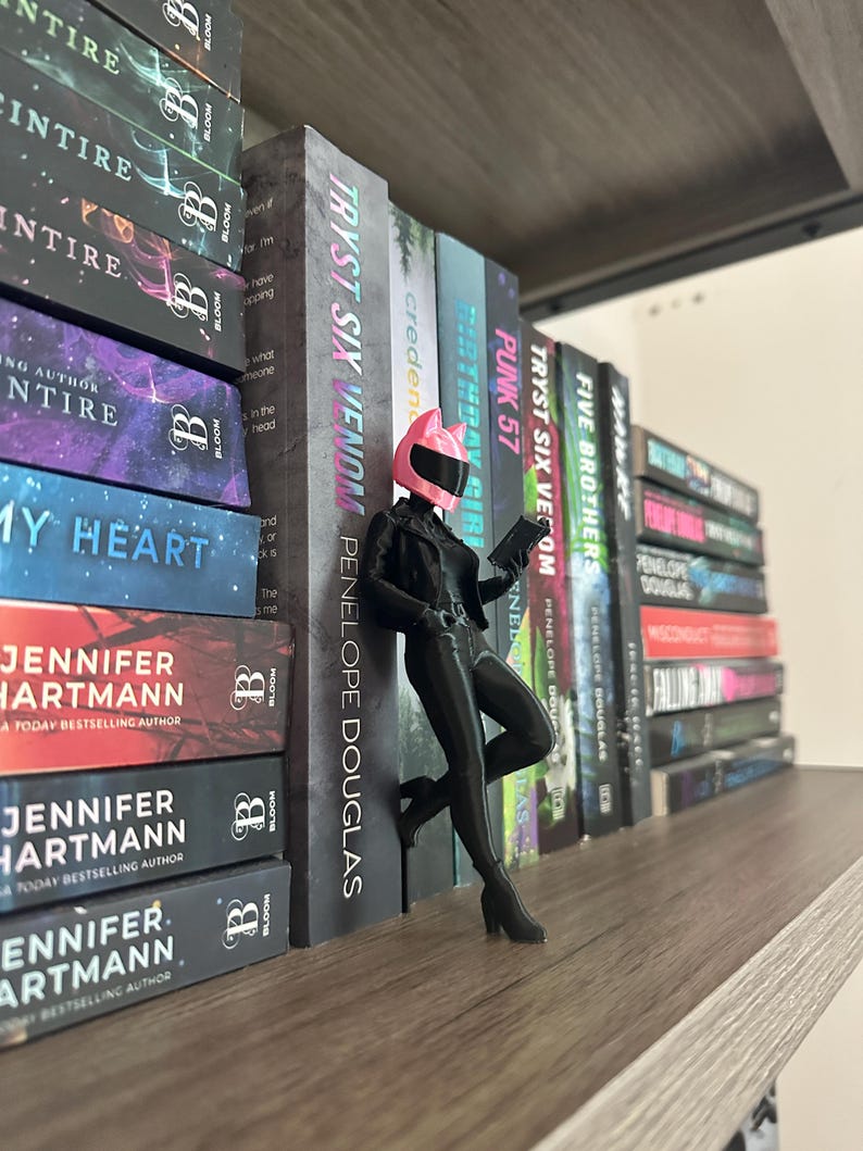 May include: A shelf displays a collection of books with titles like "TRYST SIX VENOM" and "PUNK 57." A figurine in a black outfit and pink helmet leans against the books. The books have colorful covers and the "BLOOM" logo.
