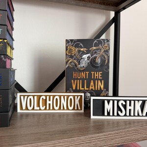Puede incluir: Una estanter&iacute;a con una pila de libros y dos placas con nombres. Las placas con nombres dicen "VOLCHONOK" y "MISHKA" en letras blancas. Tambi&eacute;n se ve un libro titulado "HUNT THE VILLAIN". Los libros est&aacute;n en una estanter&iacute;a de madera.