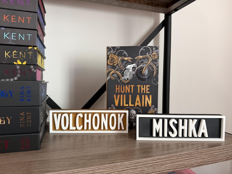Puede incluir: Una estanter&iacute;a con una pila de libros y dos placas con nombres. Las placas con nombres dicen "VOLCHONOK" y "MISHKA" en letras blancas. Tambi&eacute;n se ve un libro titulado "HUNT THE VILLAIN". Los libros est&aacute;n en una estanter&iacute;a de madera.
