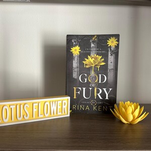 Może przedstawiać: Książka zatytułowana "God of Fury" z ż&oacute;łtym wzorem kwiatu lotosu, ż&oacute;łtym znakiem "Lotus Flower" i drukowanym w 3D ż&oacute;łtym kwiatem lotosu na p&oacute;łce. Okładka książki zawiera tekst "I'M TAKEN BY A BEAUTIFUL NIGHTMARE".
