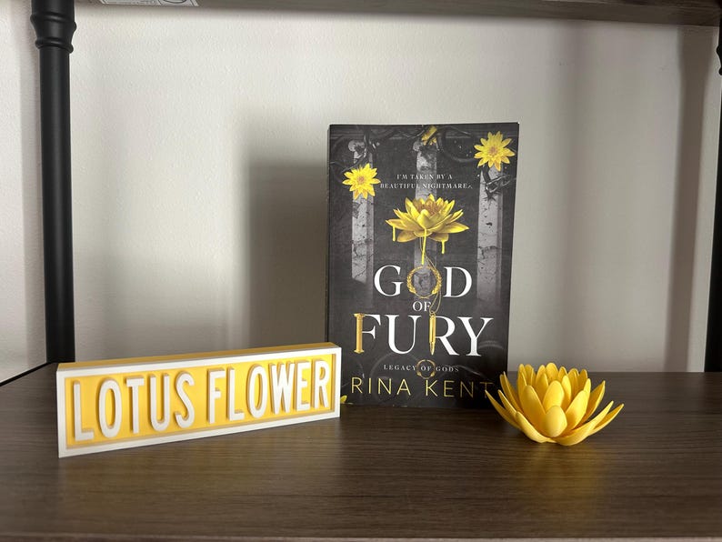 Może przedstawiać: Książka zatytułowana "God of Fury" z ż&oacute;łtym wzorem kwiatu lotosu, ż&oacute;łtym znakiem "Lotus Flower" i drukowanym w 3D ż&oacute;łtym kwiatem lotosu na p&oacute;łce. Okładka książki zawiera tekst "I'M TAKEN BY A BEAUTIFUL NIGHTMARE".