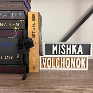 Puede incluir: Una pila de libros de Rina Kent, una figura negra y dos placas con los nombres "MISHKA VOLCHONOK". El libro "Hunt the Villain" es visible. Las placas tienen letras blancas sobre fondos negros y beige.
