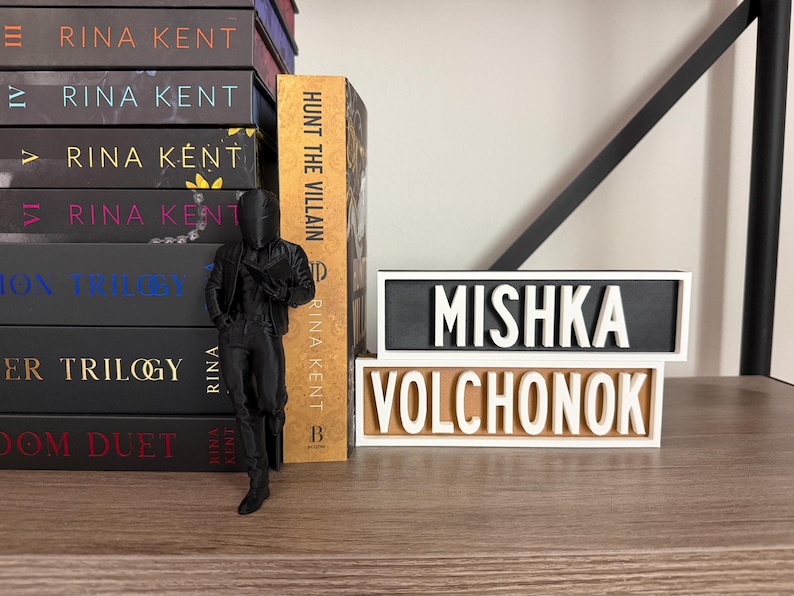 Puede incluir: Una pila de libros de Rina Kent, una figura negra y dos placas con los nombres "MISHKA VOLCHONOK". El libro "Hunt the Villain" es visible. Las placas tienen letras blancas sobre fondos negros y beige.