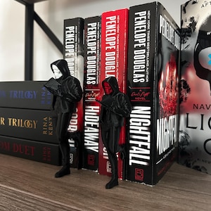 Puede incluir: Una estanter&iacute;a con libros de Penelope Douglas, con los t&iacute;tulos "Nightfall" e "Hideaway" visibles. Dos figuras negras, una con m&aacute;scara blanca y otra con m&aacute;scara roja, se encuentran frente a los libros.