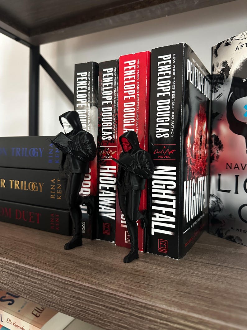 Puede incluir: Una estanter&iacute;a con libros de Penelope Douglas, con los t&iacute;tulos "Nightfall" e "Hideaway" visibles. Dos figuras negras, una con m&aacute;scara blanca y otra con m&aacute;scara roja, se encuentran frente a los libros.