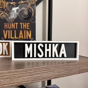 Puede incluir: Un letrero enmarcado en blanco con el nombre "MISHKA" en letras blancas y en negrita sobre un fondo negro. El letrero se muestra en una estanter&iacute;a de madera, con un libro titulado "Hunt the Villain" visible en el fondo.