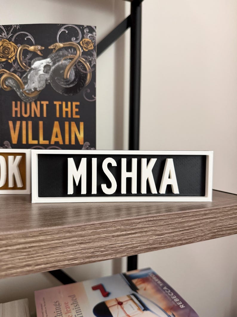 Puede incluir: Un letrero enmarcado en blanco con el nombre "MISHKA" en letras blancas y en negrita sobre un fondo negro. El letrero se muestra en una estanter&iacute;a de madera, con un libro titulado "Hunt the Villain" visible en el fondo.