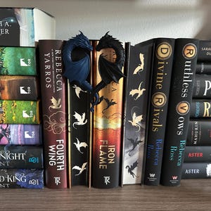 May include: A bookshelf with a variety of fantasy novels, including "Iron Flame", "Fourth Wing", "Ruthless Crows", and "Divine Rivals".  The books are arranged in a row, with a pair of blue and black dragon bookends in the center.