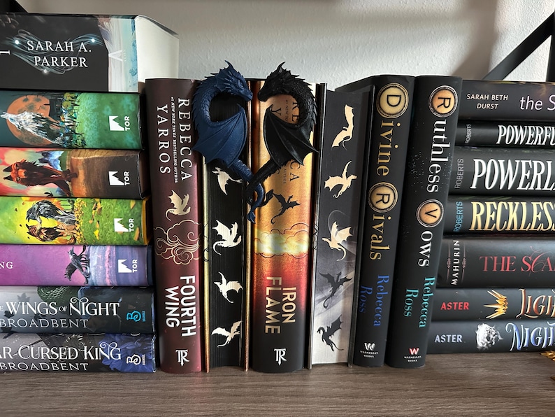 May include: A bookshelf with a variety of fantasy novels, including "Iron Flame", "Fourth Wing", "Ruthless Crows", and "Divine Rivals".  The books are arranged in a row, with a pair of blue and black dragon bookends in the center.