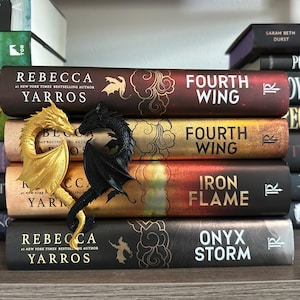 May include: A stack of five fantasy novels with dragon bookends. The books are titled "Fourth Wing", "Fourth Wing", "Iron Flame", and "Onyx Storm". The bookends are a gold and black dragon.