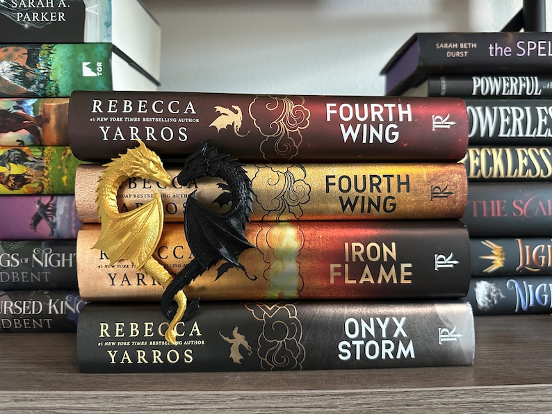 May include: A stack of five fantasy novels with dragon bookends. The books are titled "Fourth Wing", "Fourth Wing", "Iron Flame", and "Onyx Storm". The bookends are a gold and black dragon.