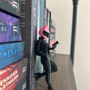 May include: A figurine of a person in a black outfit and a pink helmet, leaning against a stack of books. The figurine is holding a small device and is posed as if reading. The books have titles like "TRYST SIX VENOM" and "HEART".