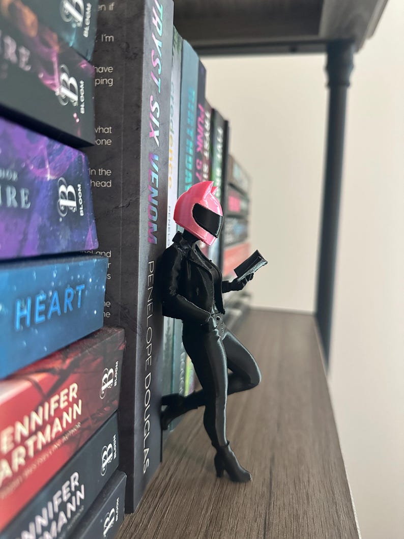 May include: A figurine of a person in a black outfit and a pink helmet, leaning against a stack of books. The figurine is holding a small device and is posed as if reading. The books have titles like "TRYST SIX VENOM" and "HEART".