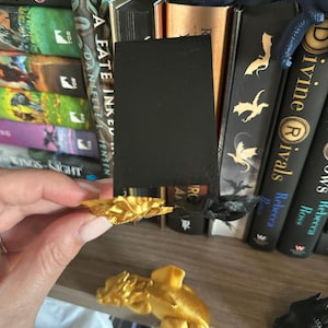 May include: Two gold dragon bookends on a bookshelf with a black bookmark. The bookshelf is filled with books with titles like "A Fate Inked" and "Divine Rivals".