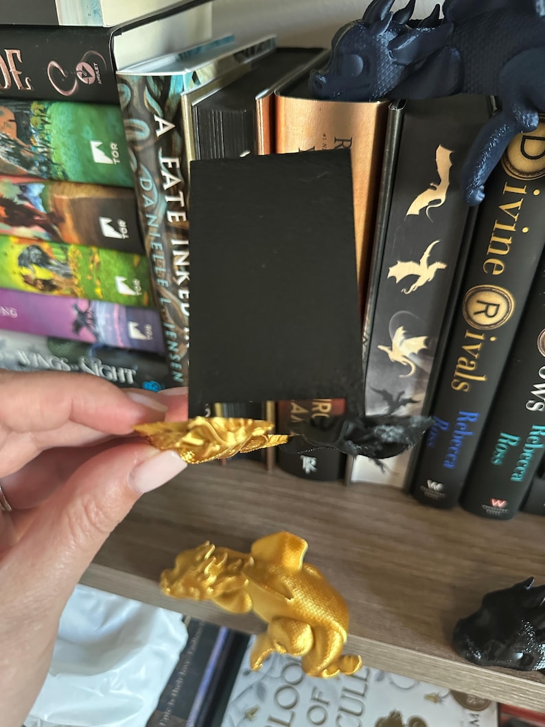 May include: Two gold dragon bookends on a bookshelf with a black bookmark. The bookshelf is filled with books with titles like "A Fate Inked" and "Divine Rivals".