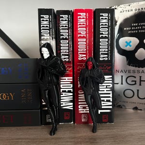 Puede incluir: Dos figuras negras de personajes con capas con capucha, una con una m&aacute;scara de calavera blanca y la otra con una m&aacute;scara roja, est&aacute;n de pie frente a una fila de libros. Los libros son de Penelope Douglas, con t&iacute;tulos como "Nightfall" e "Hideaway".