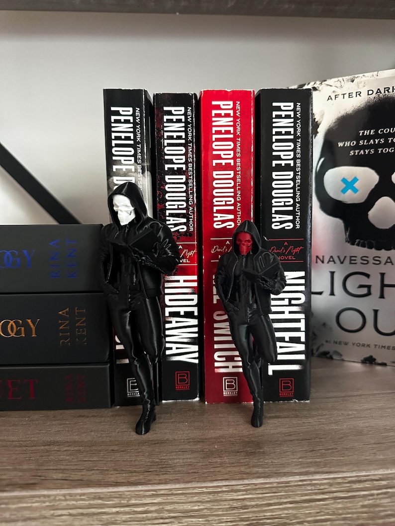 Puede incluir: Dos figuras negras de personajes con capas con capucha, una con una m&aacute;scara de calavera blanca y la otra con una m&aacute;scara roja, est&aacute;n de pie frente a una fila de libros. Los libros son de Penelope Douglas, con t&iacute;tulos como "Nightfall" e "Hideaway".