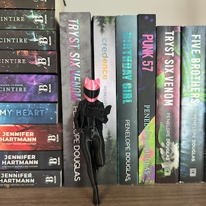 May include: A black figurine of a person wearing a pink helmet and holding a book stands in front of a shelf of books. The books have titles like "Tryst Six Venom" and "Birthday Girl". The figurine is on a wooden shelf.