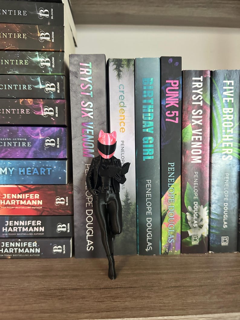 May include: A black figurine of a person wearing a pink helmet and holding a book stands in front of a shelf of books. The books have titles like "Tryst Six Venom" and "Birthday Girl". The figurine is on a wooden shelf.