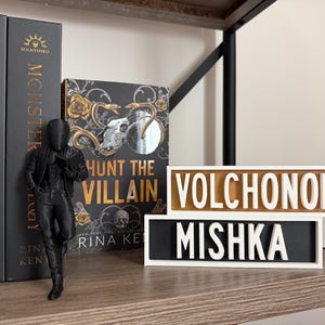 Puede incluir: Una figura negra de una persona con chaqueta y pantal&oacute;n est&aacute; junto a libros y letreros decorativos. Los libros se titulan "Deception Trilogy" y "Monster". El letrero dice "Volchonok" y "Mishka".