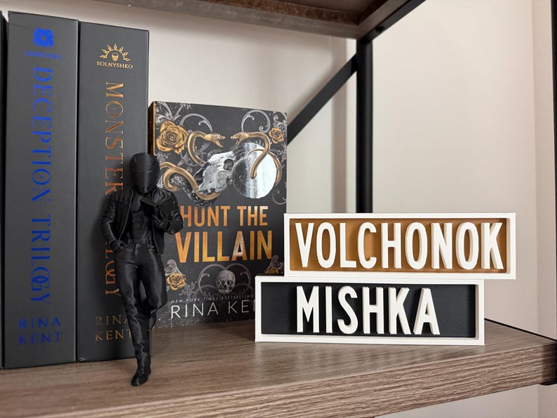 Puede incluir: Una figura negra de una persona con chaqueta y pantal&oacute;n est&aacute; junto a libros y letreros decorativos. Los libros se titulan "Deception Trilogy" y "Monster". El letrero dice "Volchonok" y "Mishka".