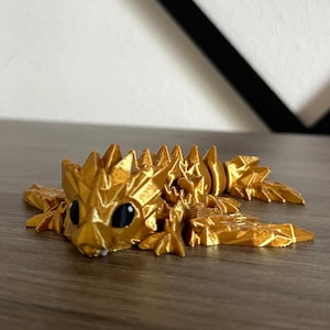 May include: A gold-colored, 3D printed, articulated, lizard-like creature with black eyes. The creature has a spiky, textured surface and is posed in a crawling position.