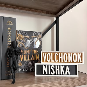 Puede incluir: Una estantería de madera con estructura metálica negra contiene una figura, libros y dos letreros rectangulares. Los letreros dicen "VOLCHONOK" y "MISHKA" en letras blancas sobre un fondo negro y dorado. Una portada de libro dice "Hunt the Villain".